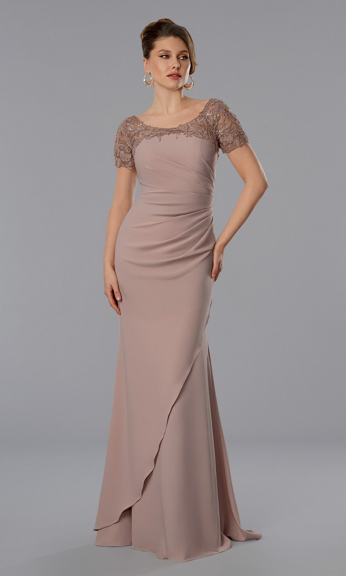 A woman wearing the blush Stella Couture 25368 long formal dress, featuring short beaded lace sleeves, a fitted bodice, and draped details, stands against a simple gray background.